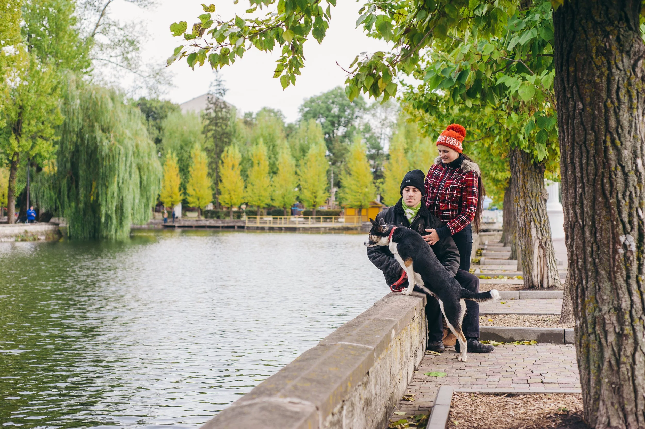 A couple with a dog standing on a stone ledge beside a river in a park with green trees and a weeping willow.