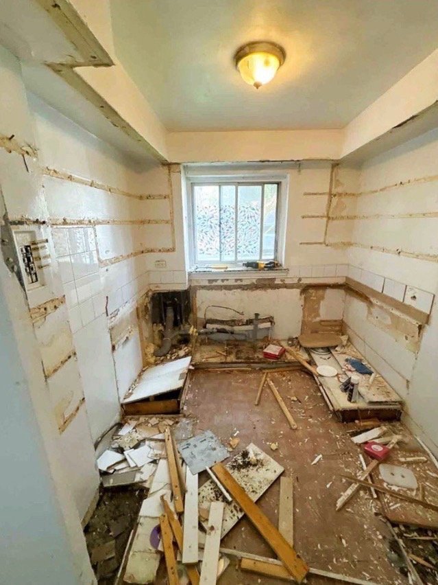 A small kitchen under renovation with damaged walls, scattered debris, and exposed plumbing, viewed from the doorway.