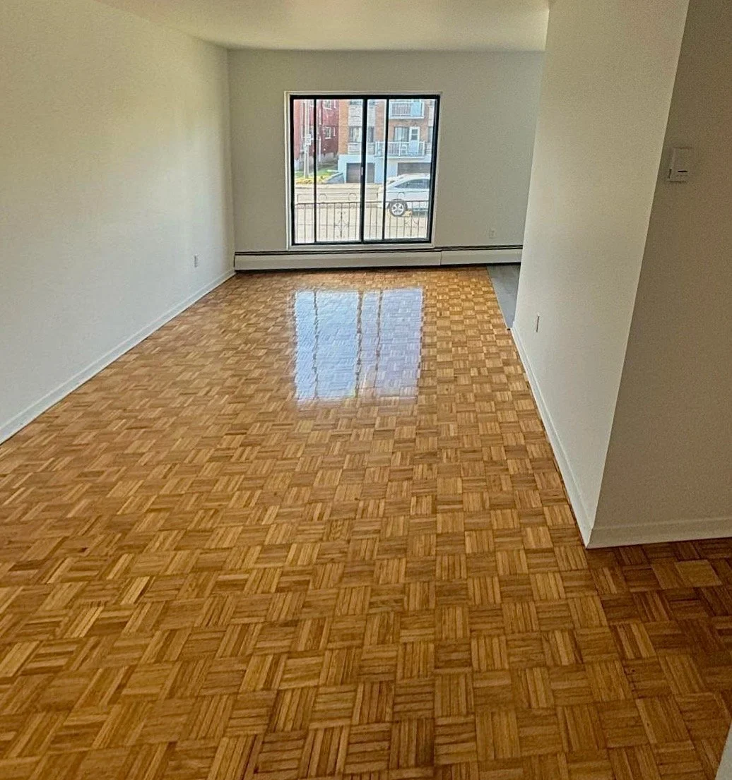 Empty room with parquet hardwood flooring, white walls, large window with a view of parked cars and neighboring buildings.