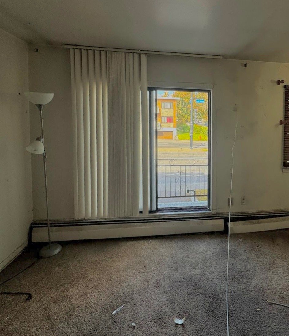 Empty living room with beige carpet, a tall floor lamp, and vertical blinds covering a sliding glass door that opens to a small balcony with a view of a street and buildings outside.