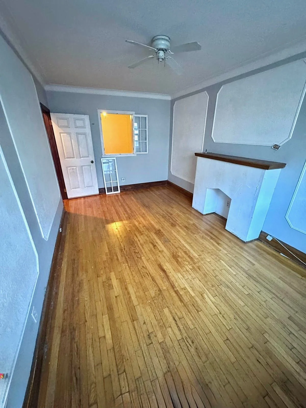 Empty room with light blue walls, hardwood floors, ceiling fan, fireplace with wooden mantel, small window, and open door with a view into another room.