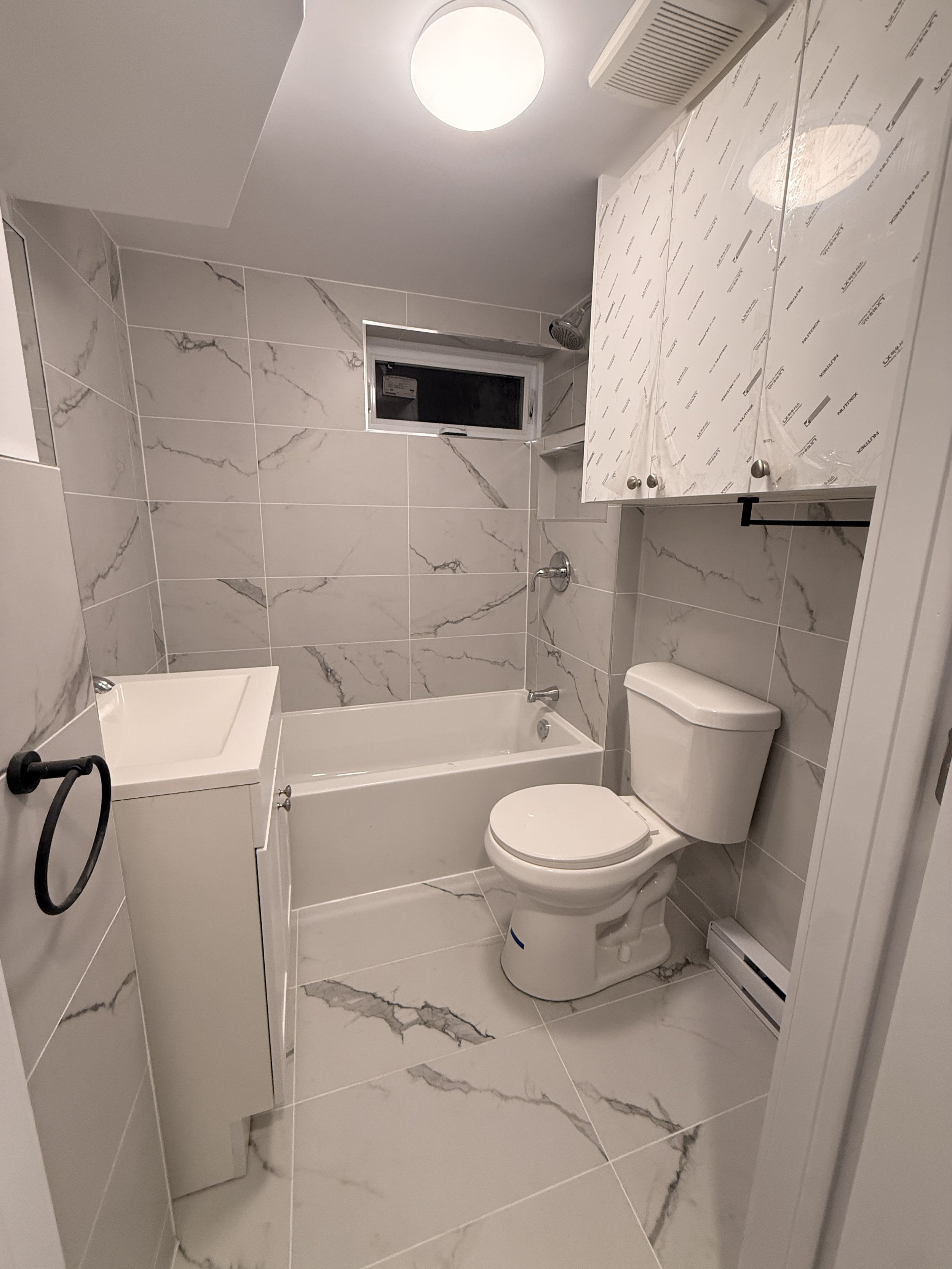 Bathroom with marble-style tiles, a bathtub, a toilet, a small sink, storage cabinets, a small window, and a ceiling light.