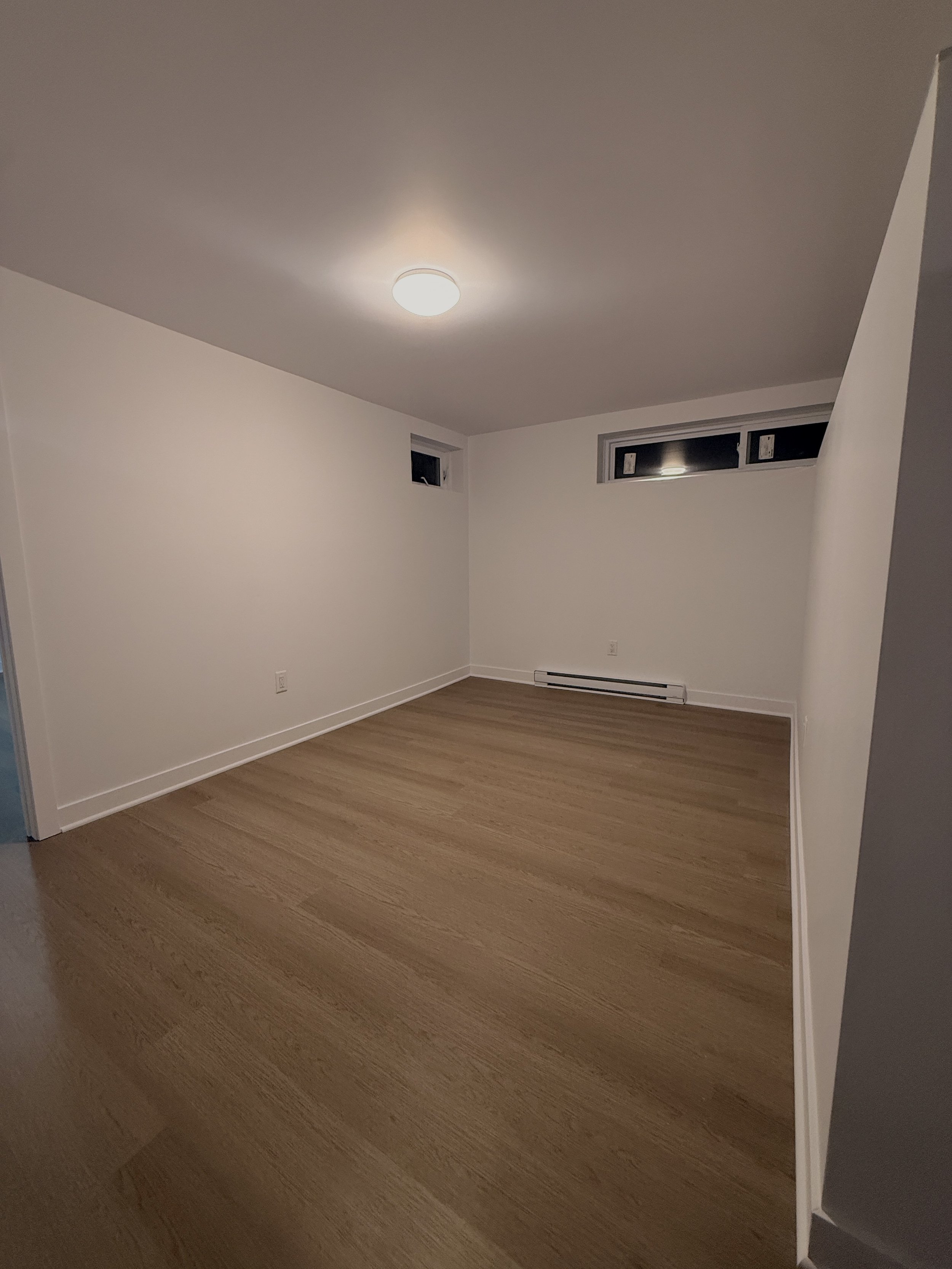 Empty room with hardwood floors, white walls, a ceiling light, and two small horizontal windows.