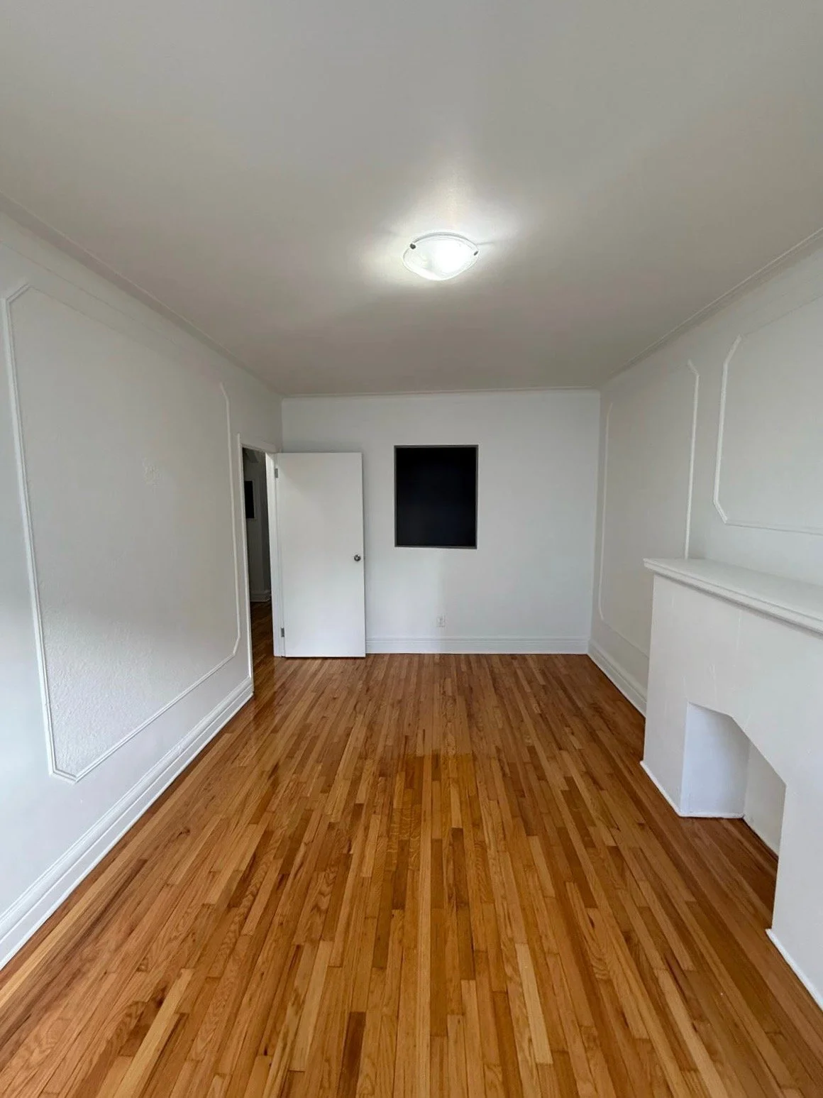 Empty living room with white walls, a hardwood floor, a ceiling light, a small open door, and a fireplace with a shelf, with a black rectangle on the wall at the back.