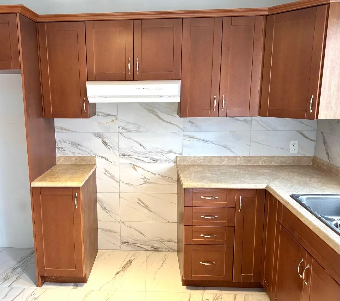 Kitchen with wooden cabinets, marble tiled wall, beige countertops, and a sink on the right.