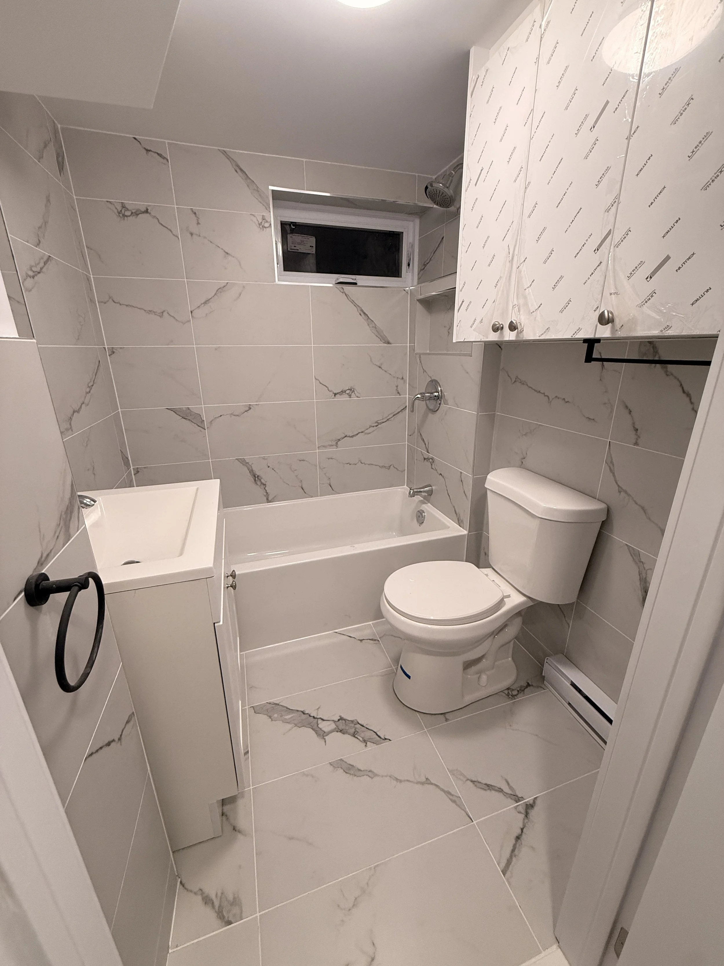 A modern bathroom with marble-patterned tiles on the walls and floor. It features a white bathtub, a white toilet, a small white utility sink, white cabinetry, and a small window.