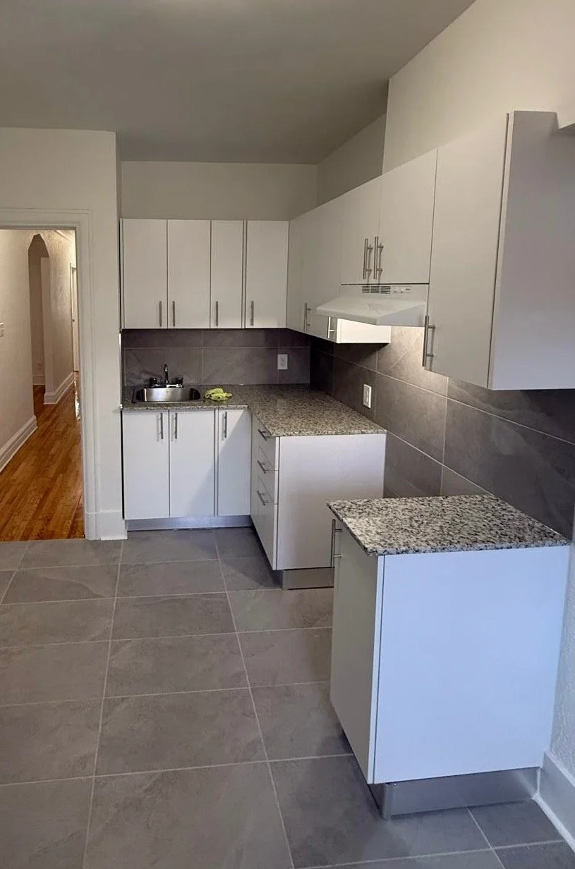 Empty kitchen with white cabinets, granite countertops, gray tiled backsplash, and gray tiled floor. Small sink with a yellow cleaning cloth, adjacent to an open doorway revealing a hallway with wooden flooring.