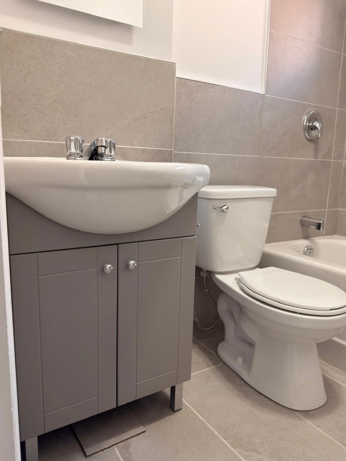 A small bathroom with a gray cabinet sink, a white toilet, and a partial view of a bathtub with a grab bar, tiled walls, and floor.