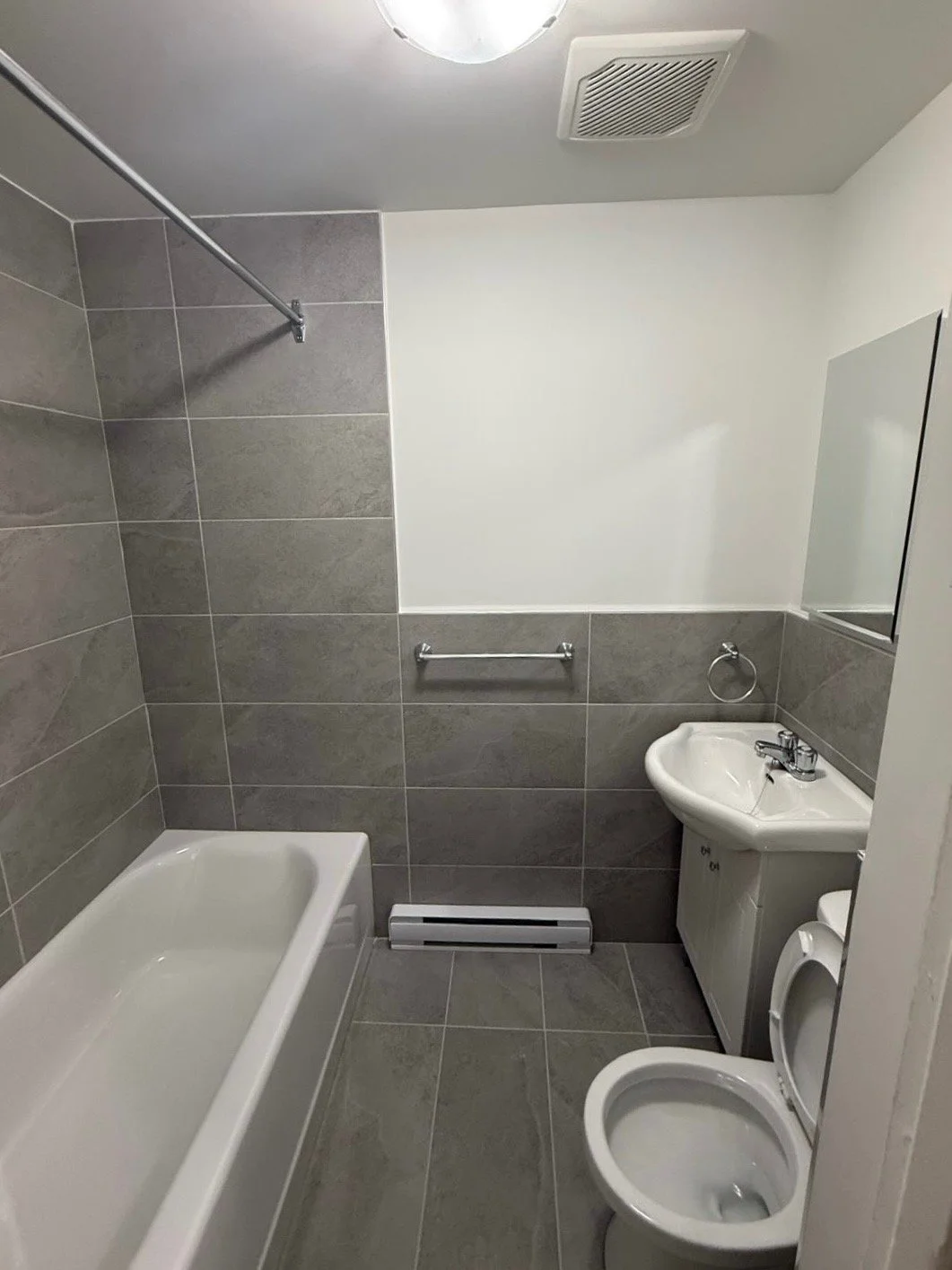 Small bathroom with gray tiled walls and floor, a white bathtub, a small white sink with cabinet, a toilet, and a mirror. There is a vent on the ceiling and a towel bar on the wall.