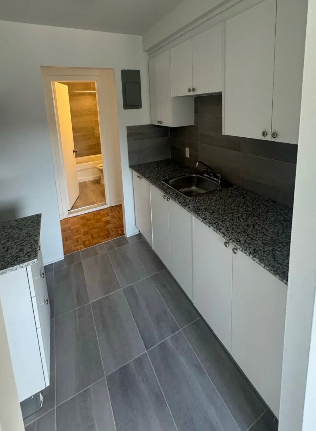 Small kitchen with white cabinets, black granite countertops, gray tile flooring, and a small sink. A doorway leads to a bathroom with a bathtub and wooden-paneled walls.