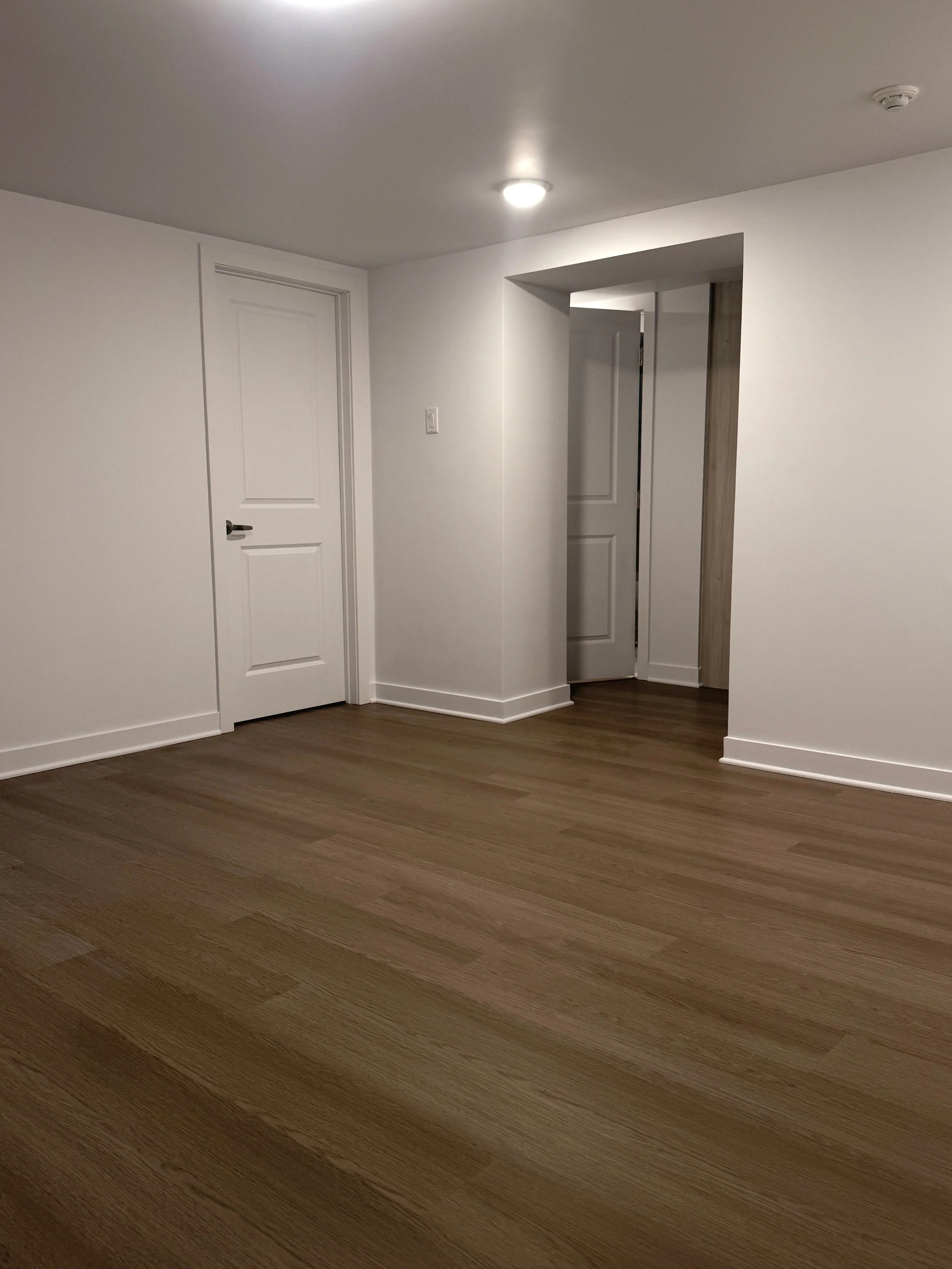 Empty room with white walls, wooden flooring, two white doors, and ceiling lights.
