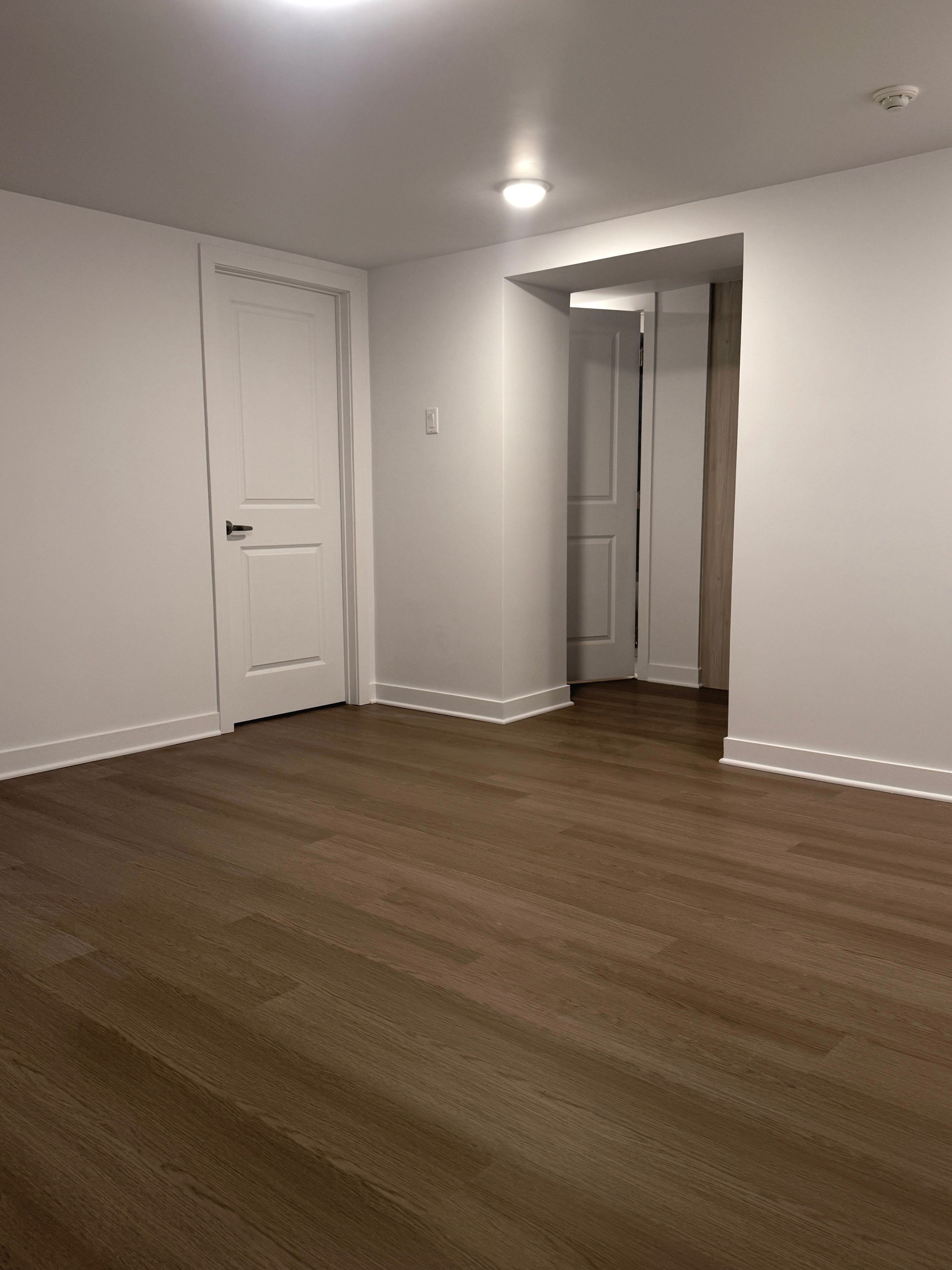 Empty room with white walls, hardwood flooring, and two closed white doors, one on the left and one in the corner.