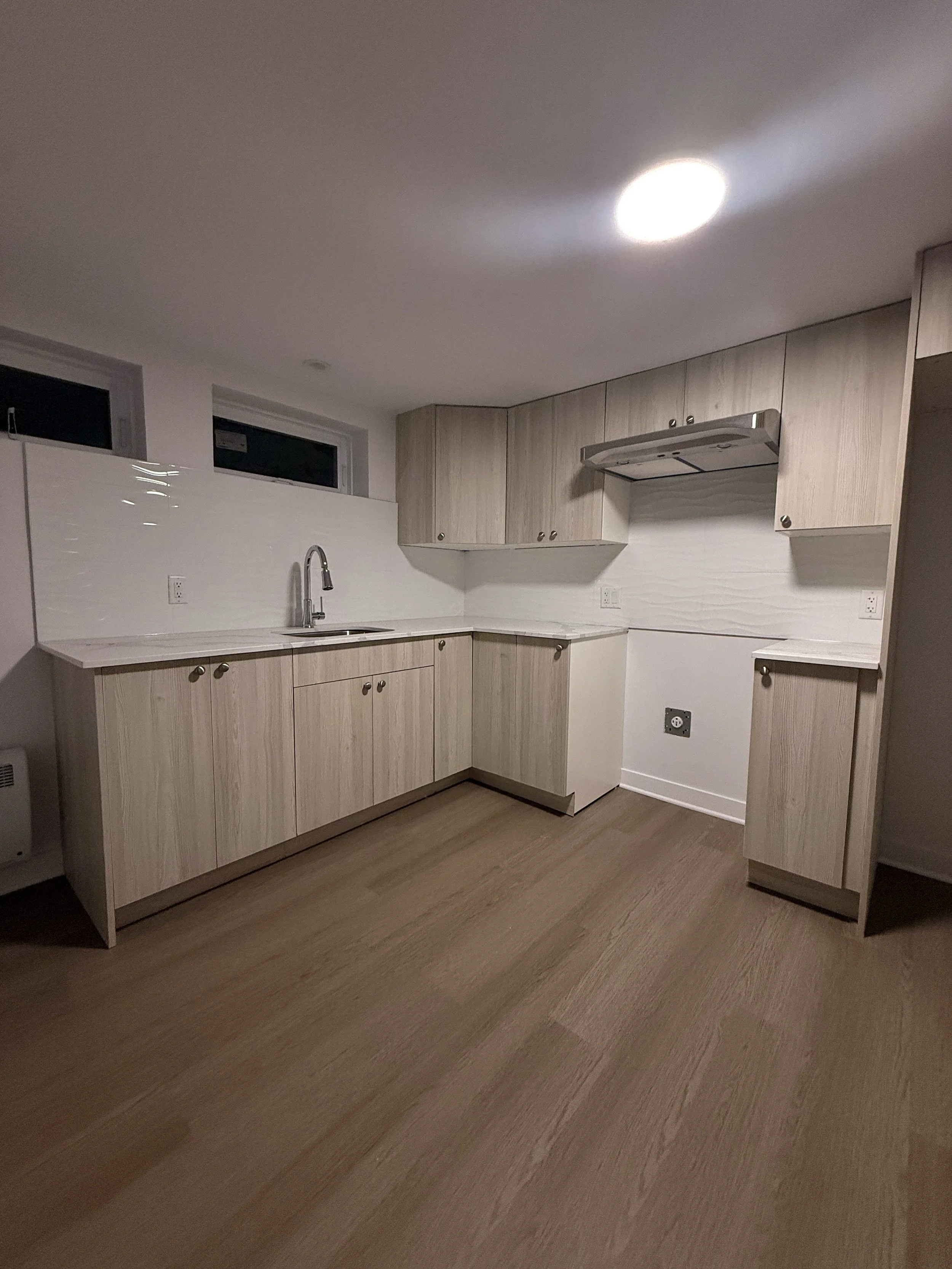 Empty kitchen with light wood cabinets, white countertops, a modern faucet, and a wood floor; small windows above the sink, and a ceiling light