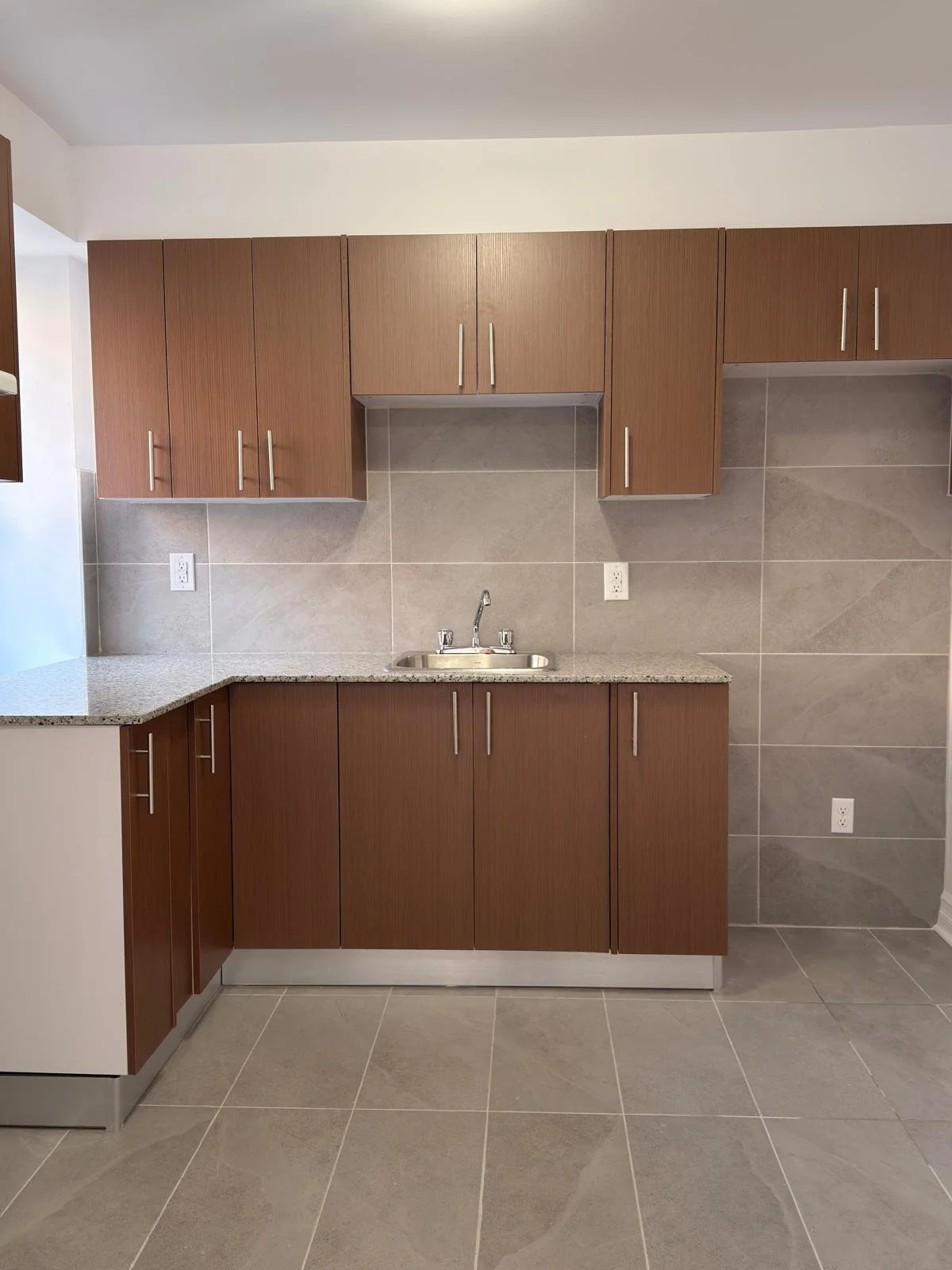 A kitchen with brown upper and lower cabinets, a small stainless steel sink with a faucet, gray tiled walls, and large gray tiled flooring.