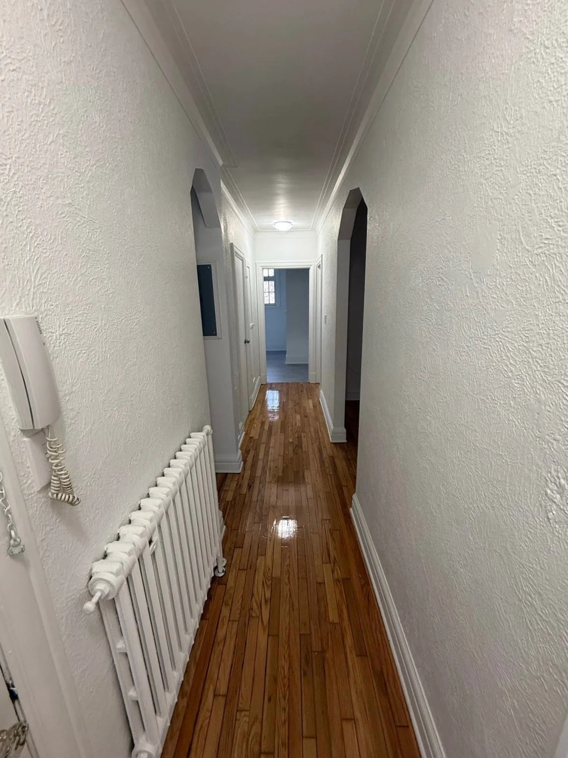 Narrow hallway with hardwood floor, white textured walls, white ceiling, and arched doorways leading to other rooms.