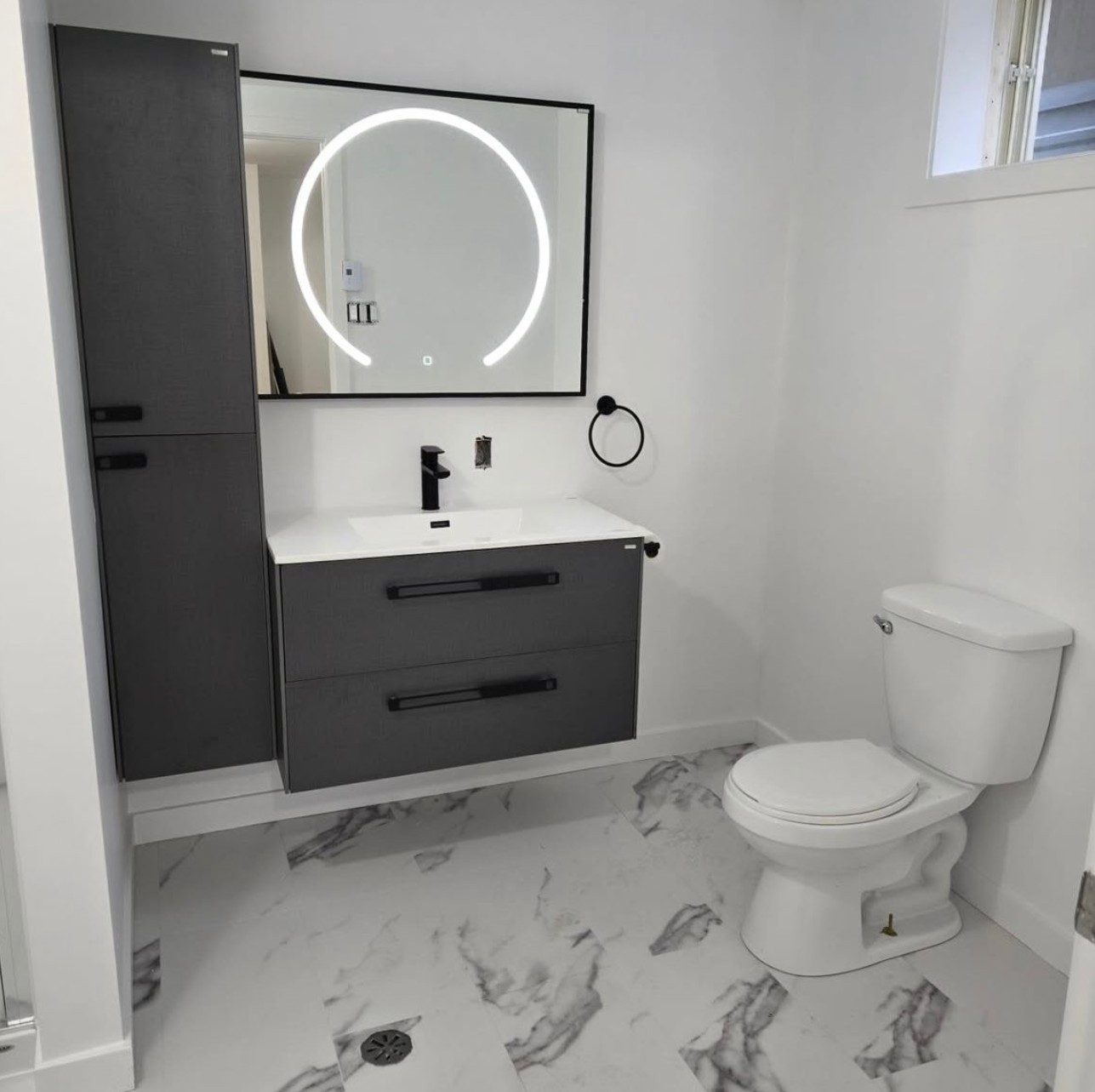 Modern bathroom with white walls, marble floor, gray vanity with drawers, a white sink, black faucet, large round mirror with LED lighting, a black towel ring, a white toilet, and a small window.