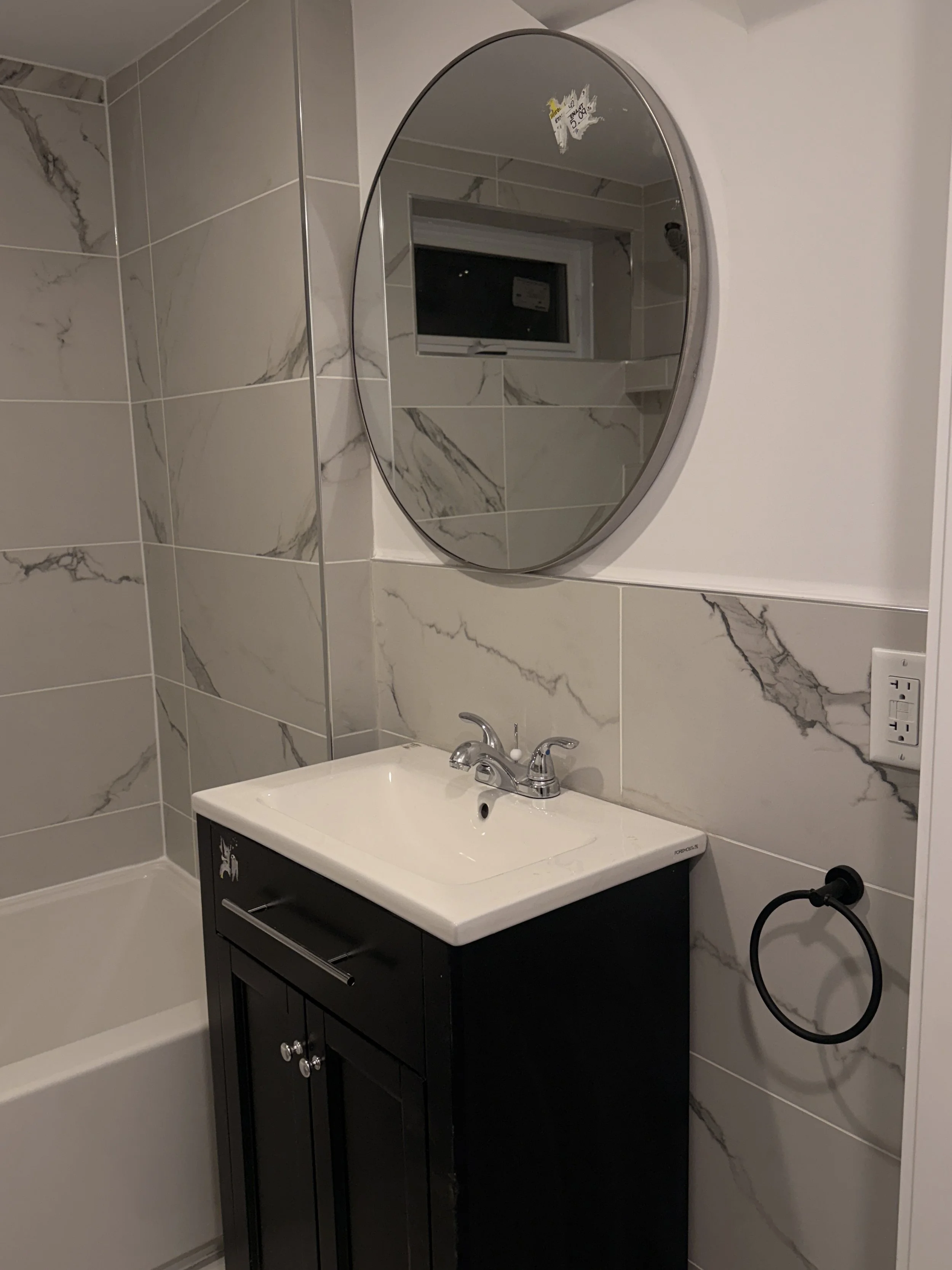Bathroom with marble-tile walls, a round mirror, a small black vanity with a white sink, a faucet, a wall socket, and a towel ring.