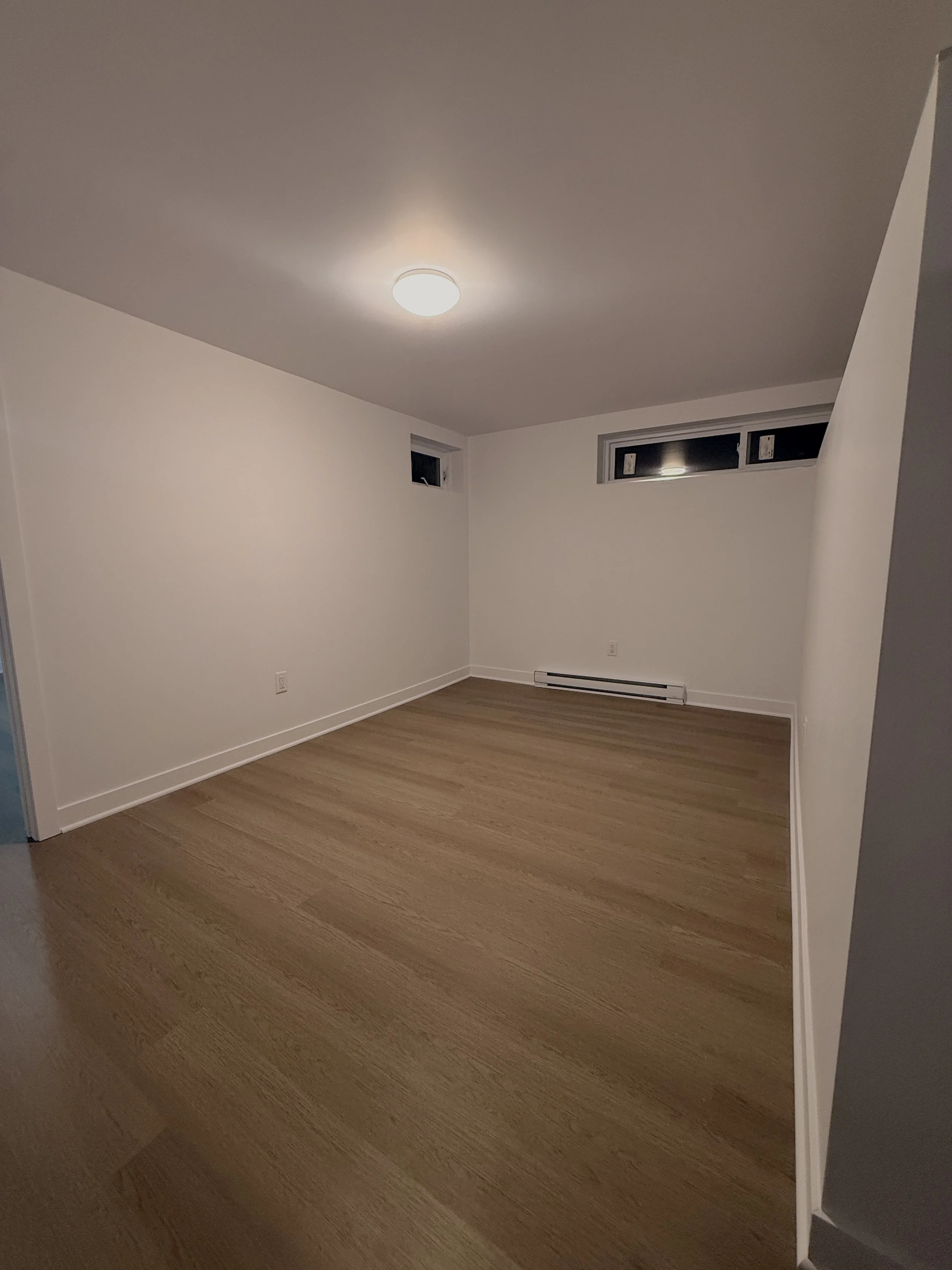 Empty room with white walls, wood flooring, small rectangular windows, and a ceiling light.