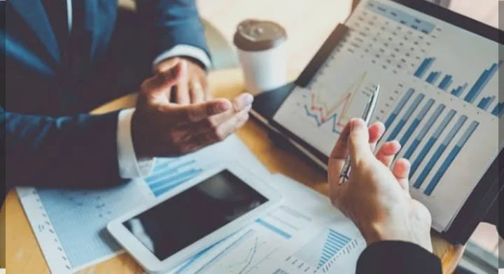 Two people analyzing financial data on digital tablets and a laptop with bar and line charts, in a business meeting setting.