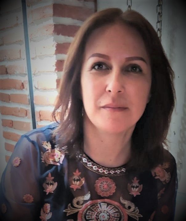 Retrato de una mujer con cabello castaño y piel clara, usando una blusa azul con bordados coloridos y un collar, en un fondo de pared de ladrillos y estructuras metálicas.