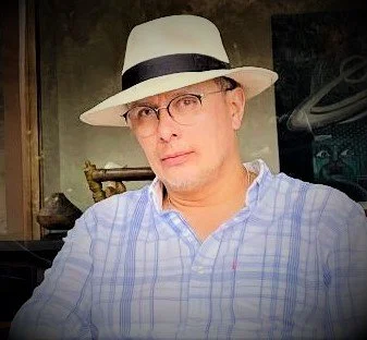 Hombre con gafas y sombrero blanco, camisa de cuadros azul y fondo oscuro.