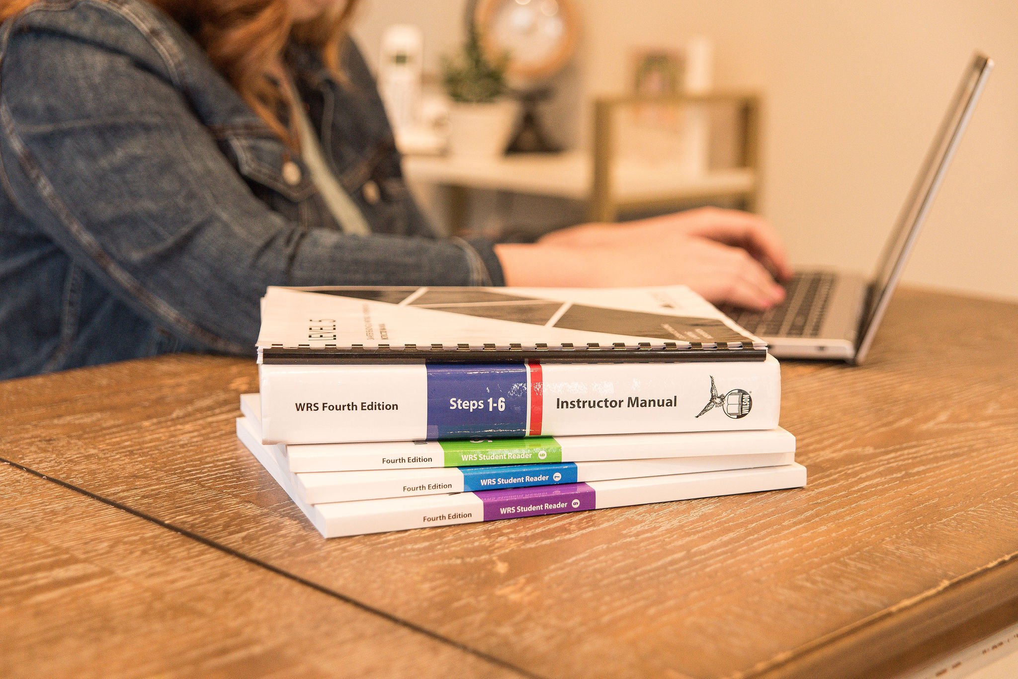 A stack of educational books about step-by-step instructional guides, with a laptop and person working in the background.