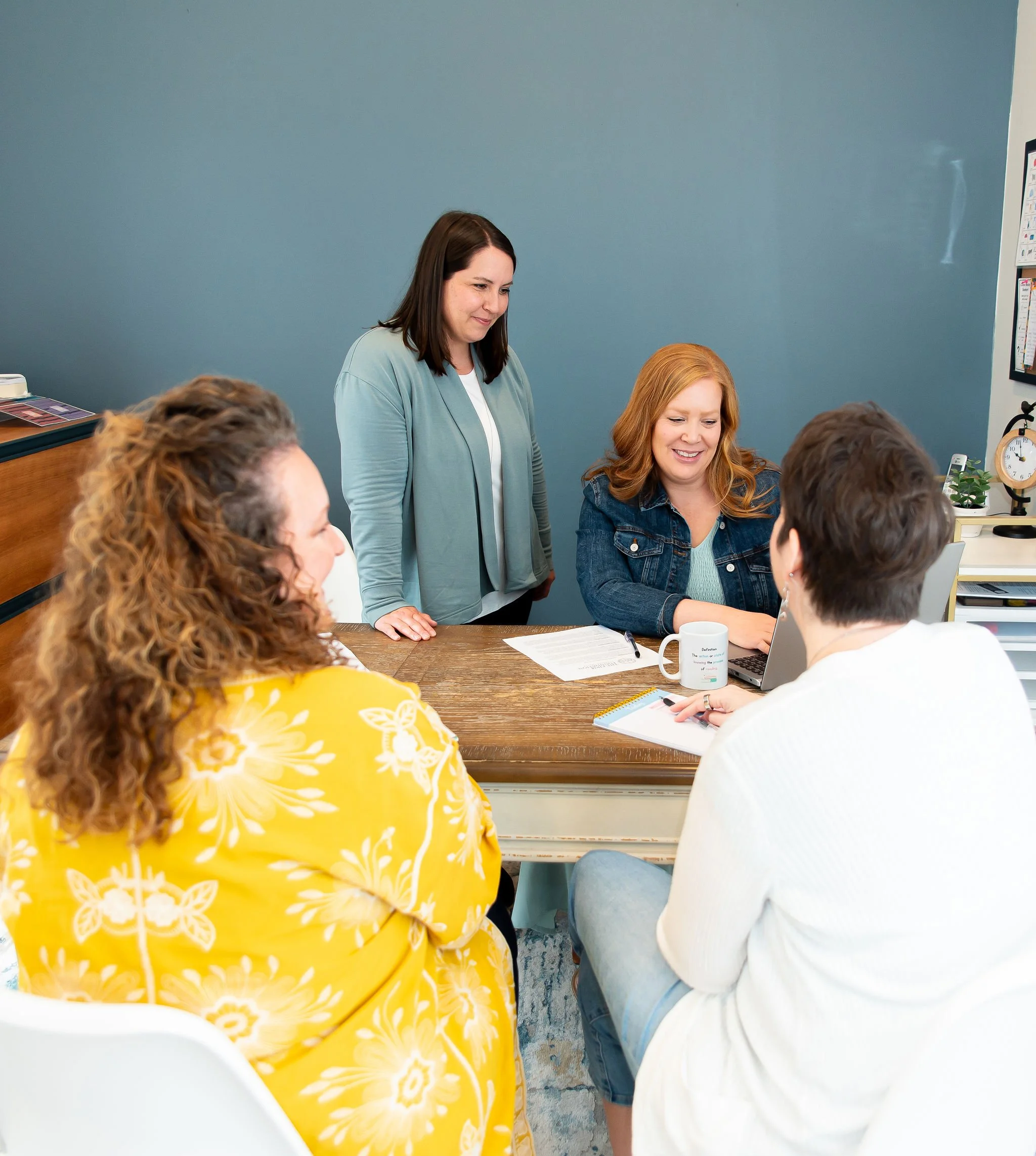 A group of people gathered around a table in a meeting room. Two women are standing and smiling, while others are sitting and engaged in a discussion or looking at a laptop. The room has a dark teal wall, and the table has various papers, notebooks, and coffee mugs.