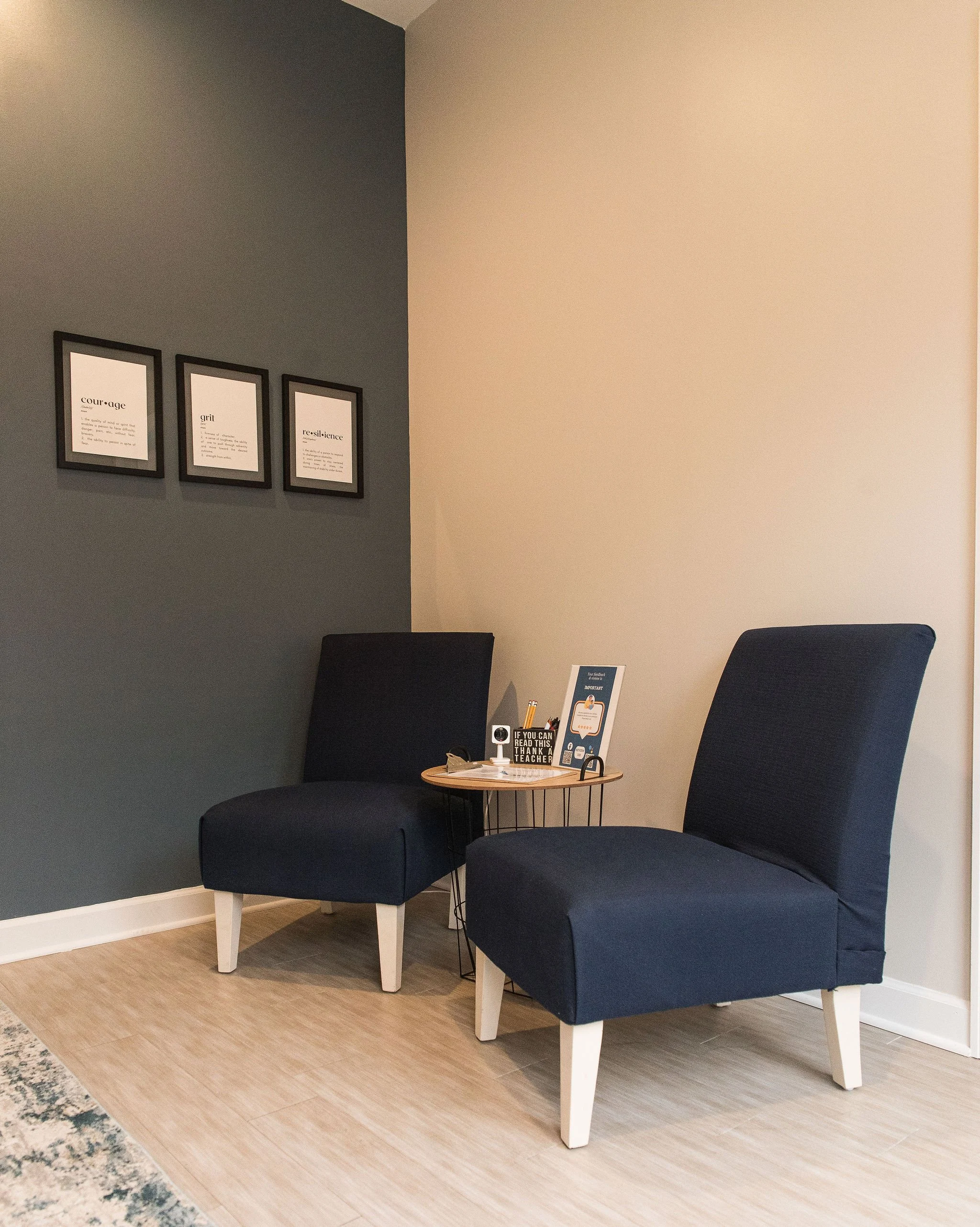 A cozy seating area with a dark blue armchair, a matching ottoman, and a small round wooden side table with framed quotes, papers, and pens. The wall on the left is painted dark gray, and the wall on the right is light beige. There are three framed quotes on the dark gray wall that read 'courage,' 'grit,' and 'resilience.' The floor is wooden, and there is a small, patterned rug partially visible at the bottom left.