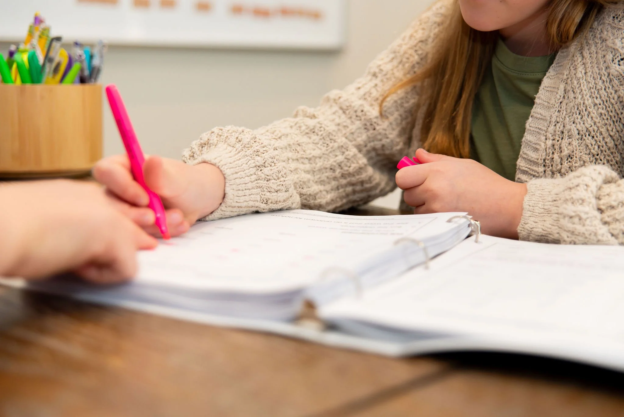 A girl with red hair, wearing a beige knitted sweater and green shirt, writing in a notebook with a pink highlighter in a classroom.