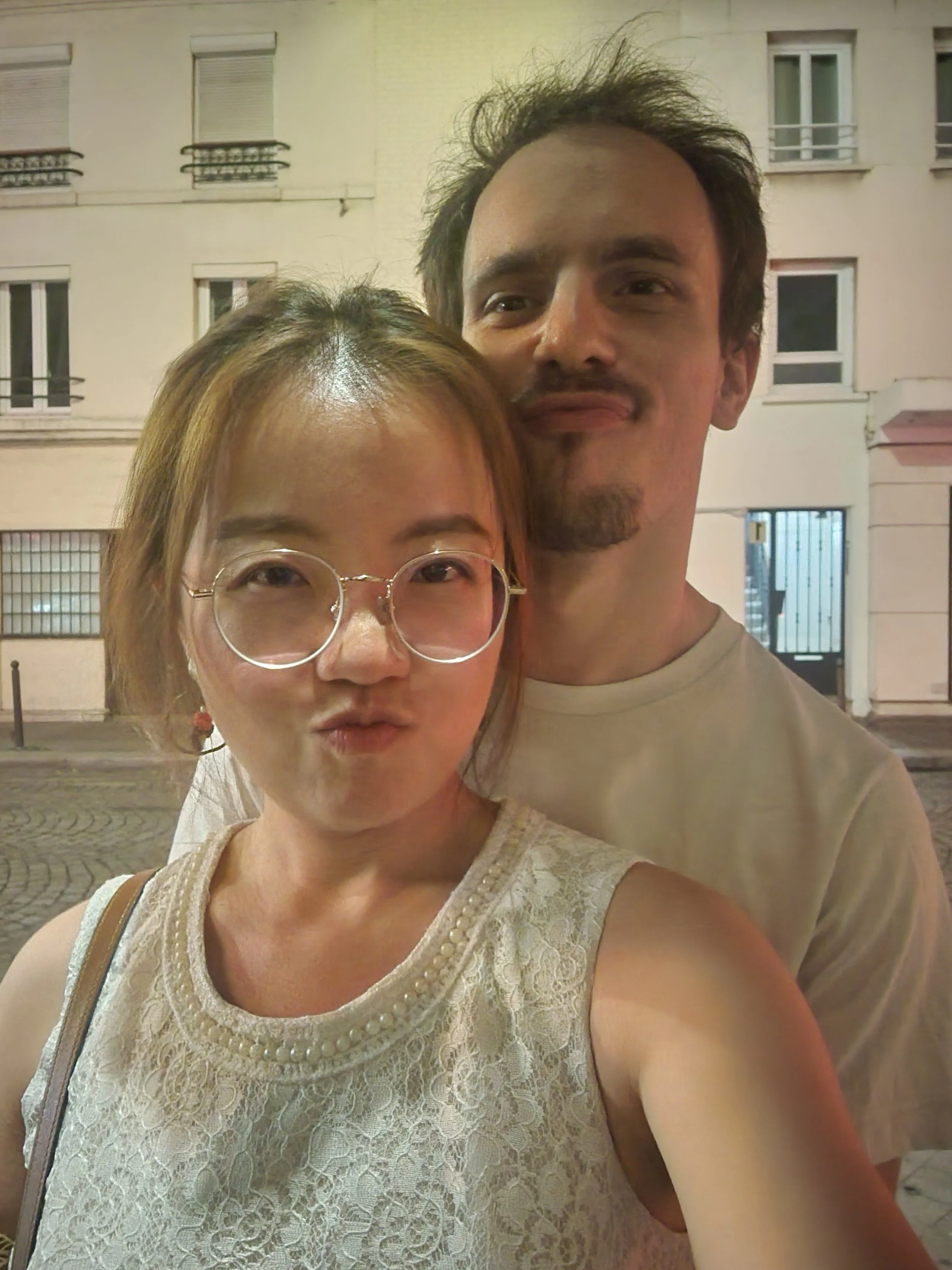Deux personnes prennent un selfie devant un bâtiment résidentiel urbain la nuit, une femme avec des lunettes et une robe en dentelle, un homme avec des cheveux noirs et une barbe, tous deux souriants.