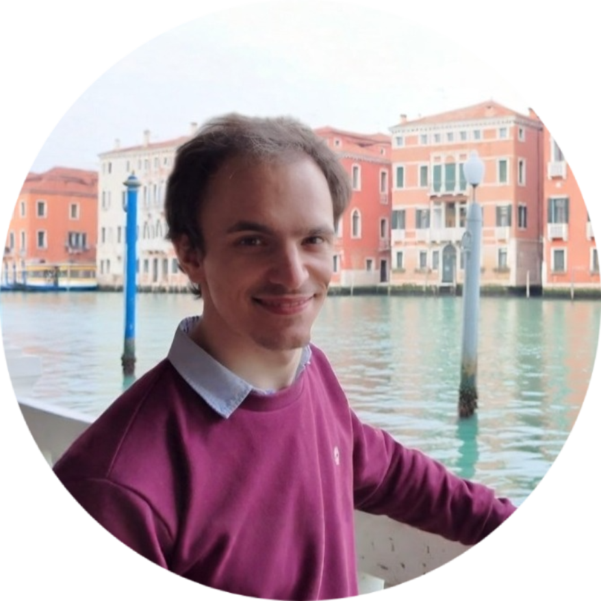 Un jeune homme souriant en manteau violet, debout près d'eau avec des bâtiments colorés en arrière-plan.