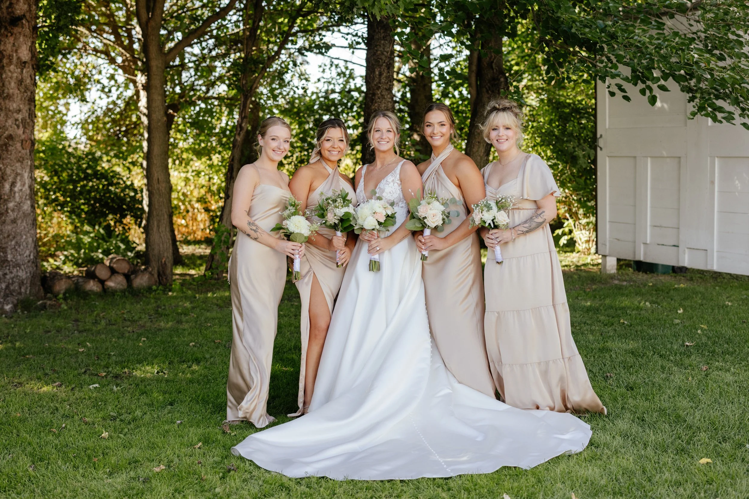 Group of five women in beige dresses holding bouquets, standing outdoors in front of trees, smiling at the camera at a daytime wedding event.