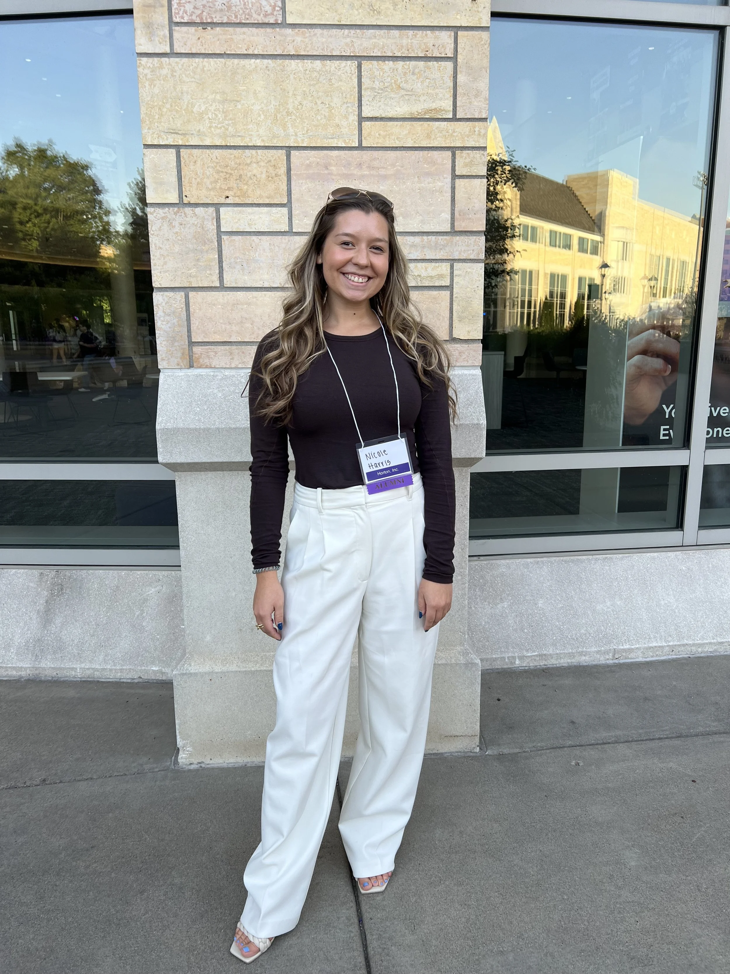 A young woman with wavy blonde hair smiling, wearing a black long-sleeve top, white pants, and open-toed shoes, standing outside a building with large glass windows and brick wall, with reflection of trees and buildings.