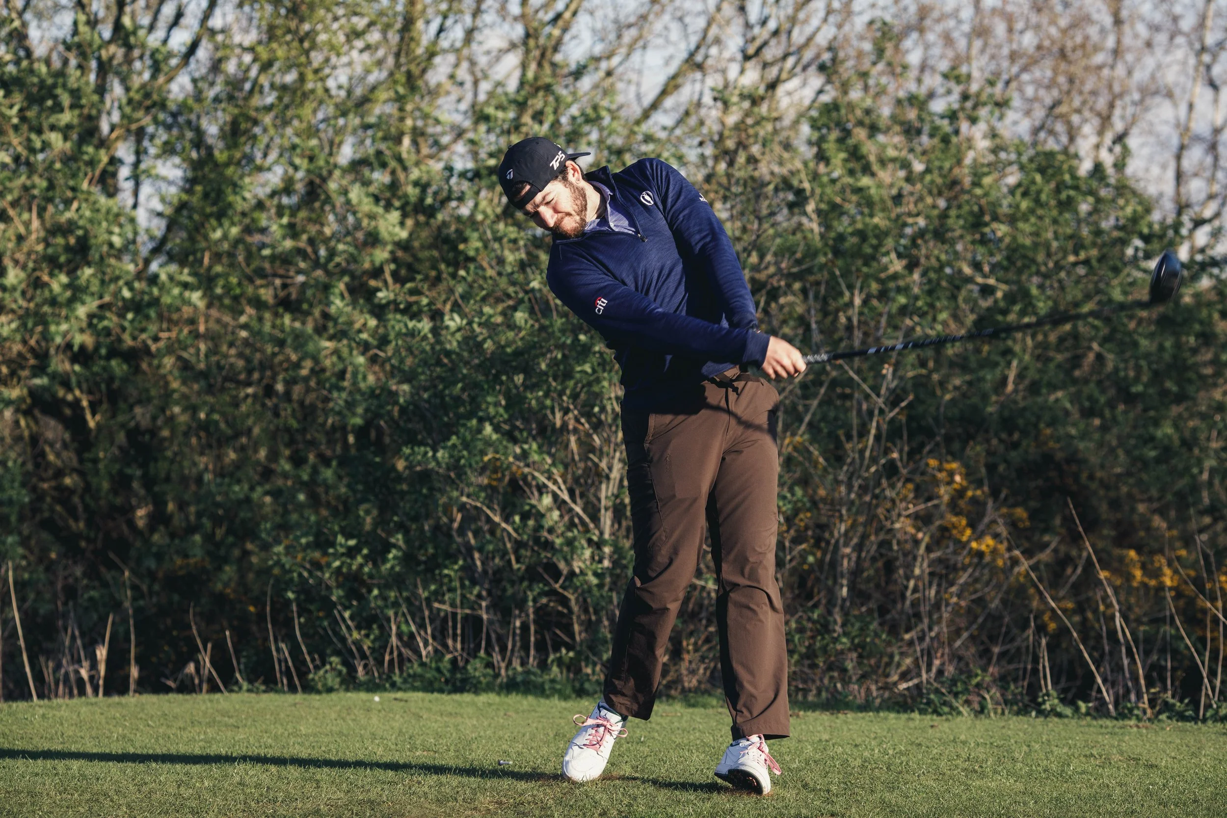 A man in a blue jacket, brown pants, and a black cap is swinging a golf club on a golf course, with trees and shrubs in the background.