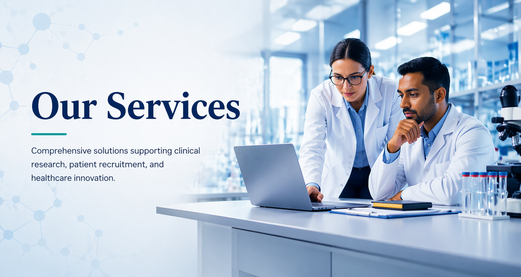 Two scientists in white lab coats working together in a laboratory, looking at a laptop screen, surrounded by scientific equipment.