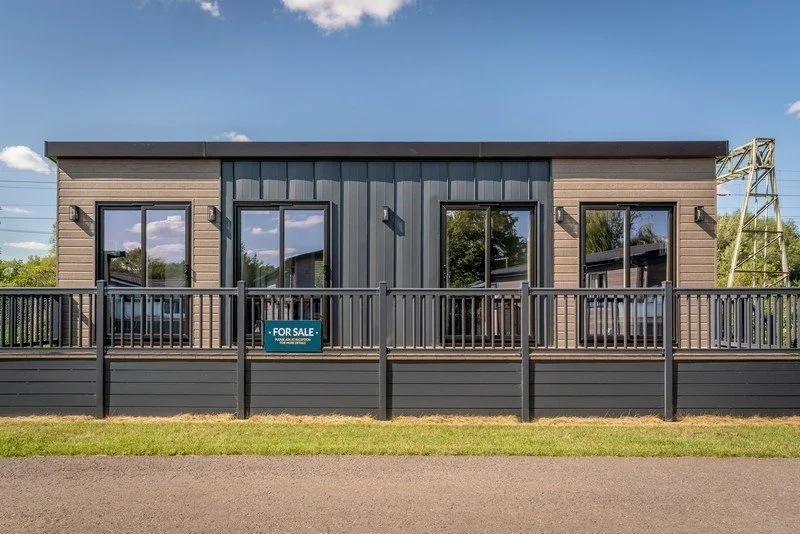 Modern two-story house with dark wood siding, large windows, and a balcony with a railing, for sale sign in front
