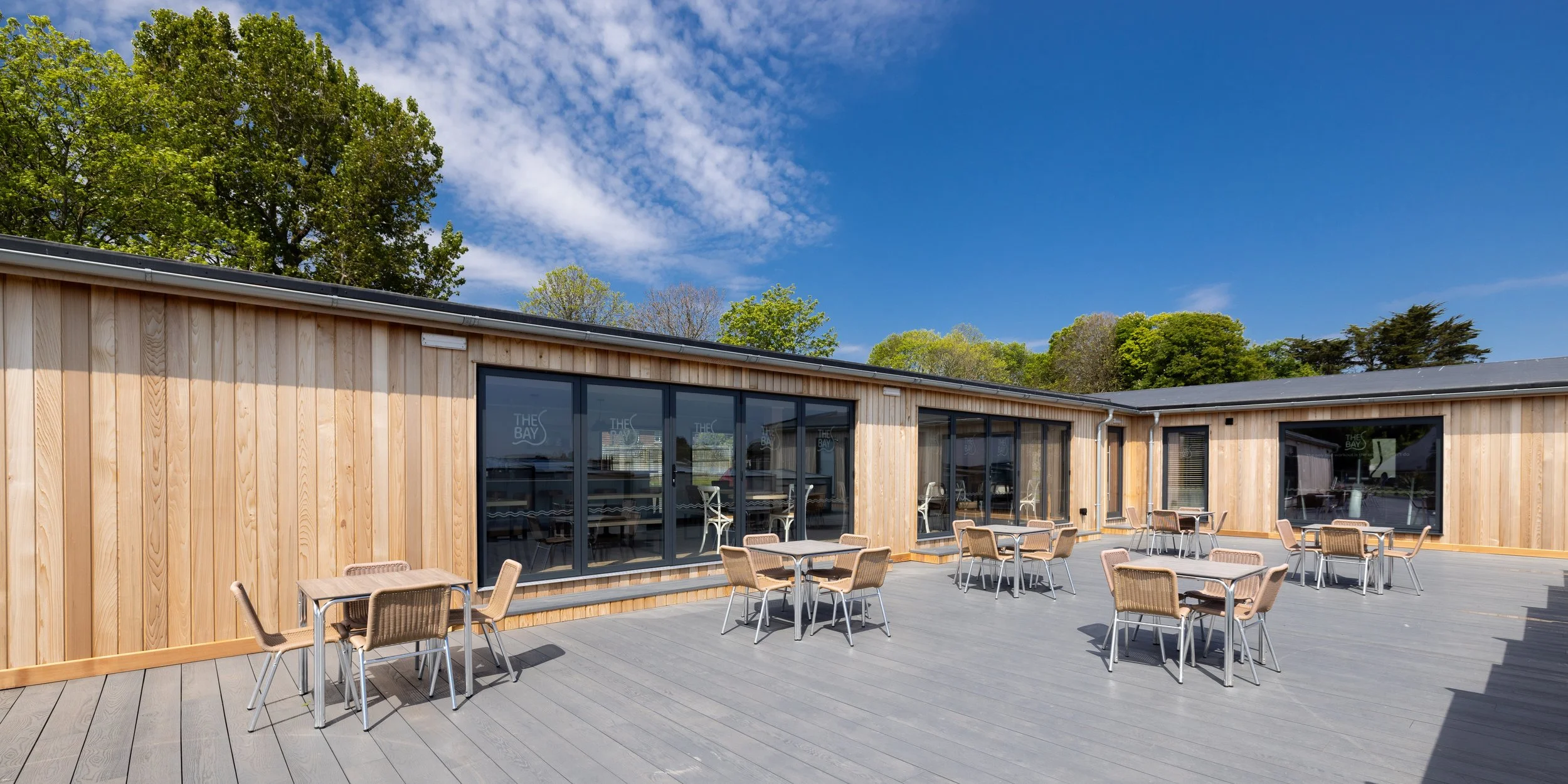 Outdoor patio with wooden building, glass windows, and tables with chairs, under a partly cloudy blue sky, surrounded by green trees.