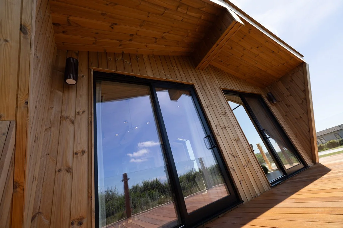 Exterior view of a modern wooden house with large glass sliding doors and a wooden deck, under a partly cloudy sky.