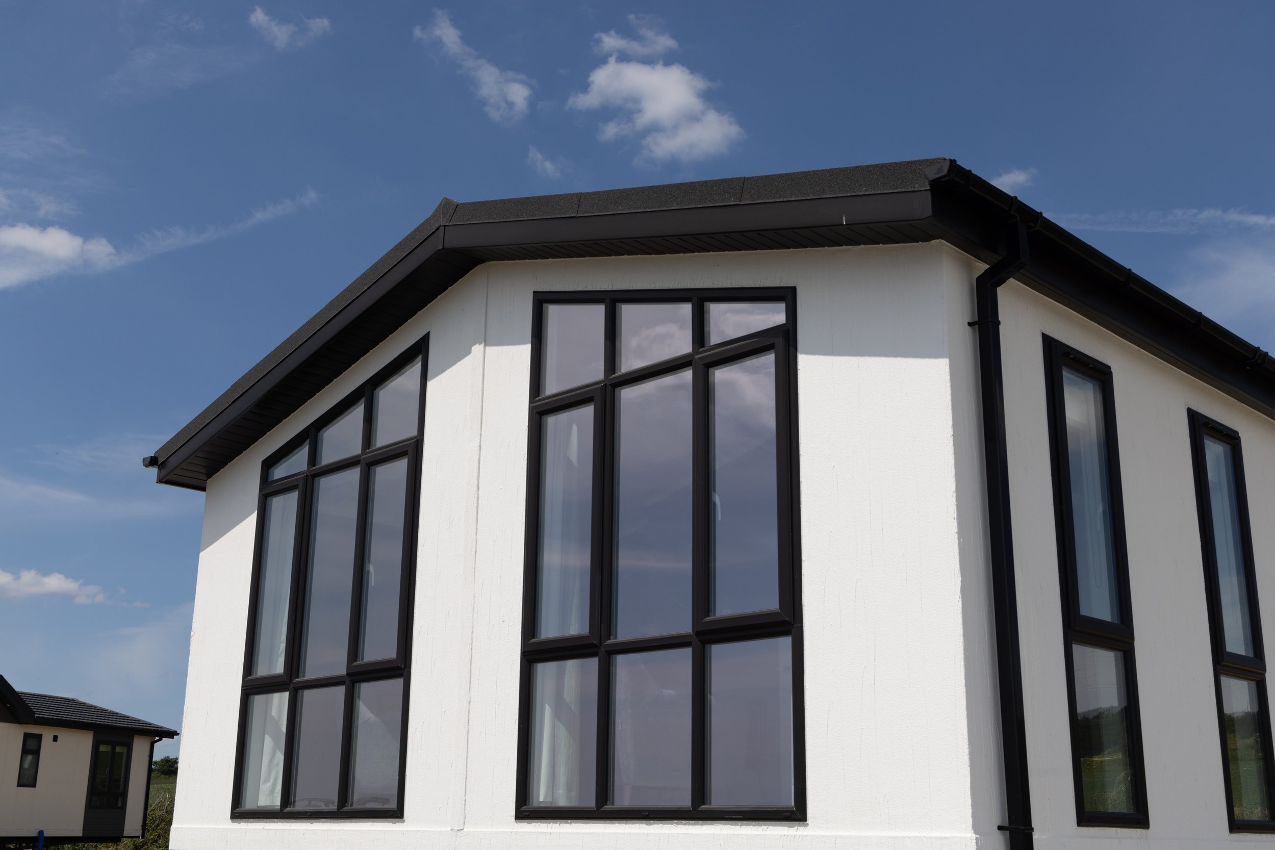 Modern white house with large black-framed windows, sloped roof, and black gutter system under a partly cloudy blue sky.