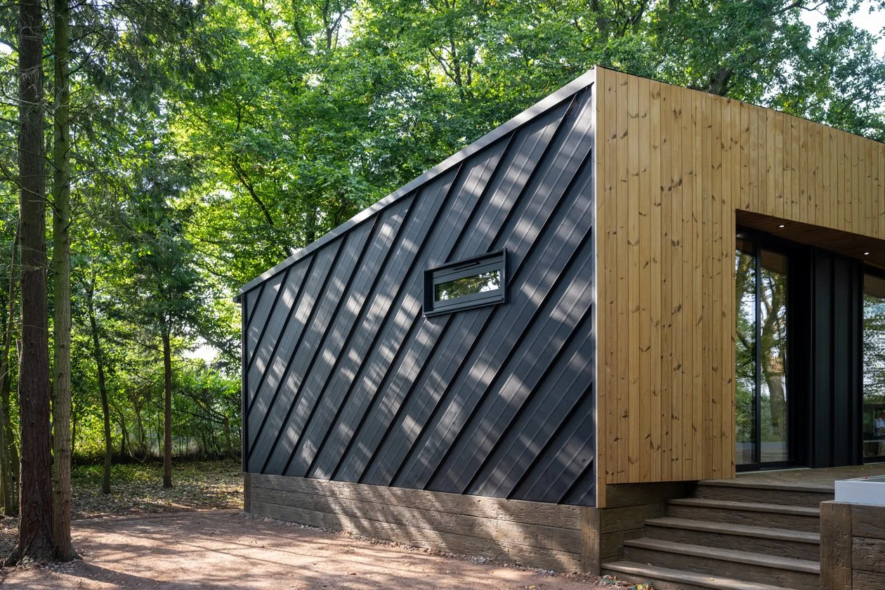 Modern house with black metal siding and wooden exterior, located in a wooded area with trees and a dirt ground.