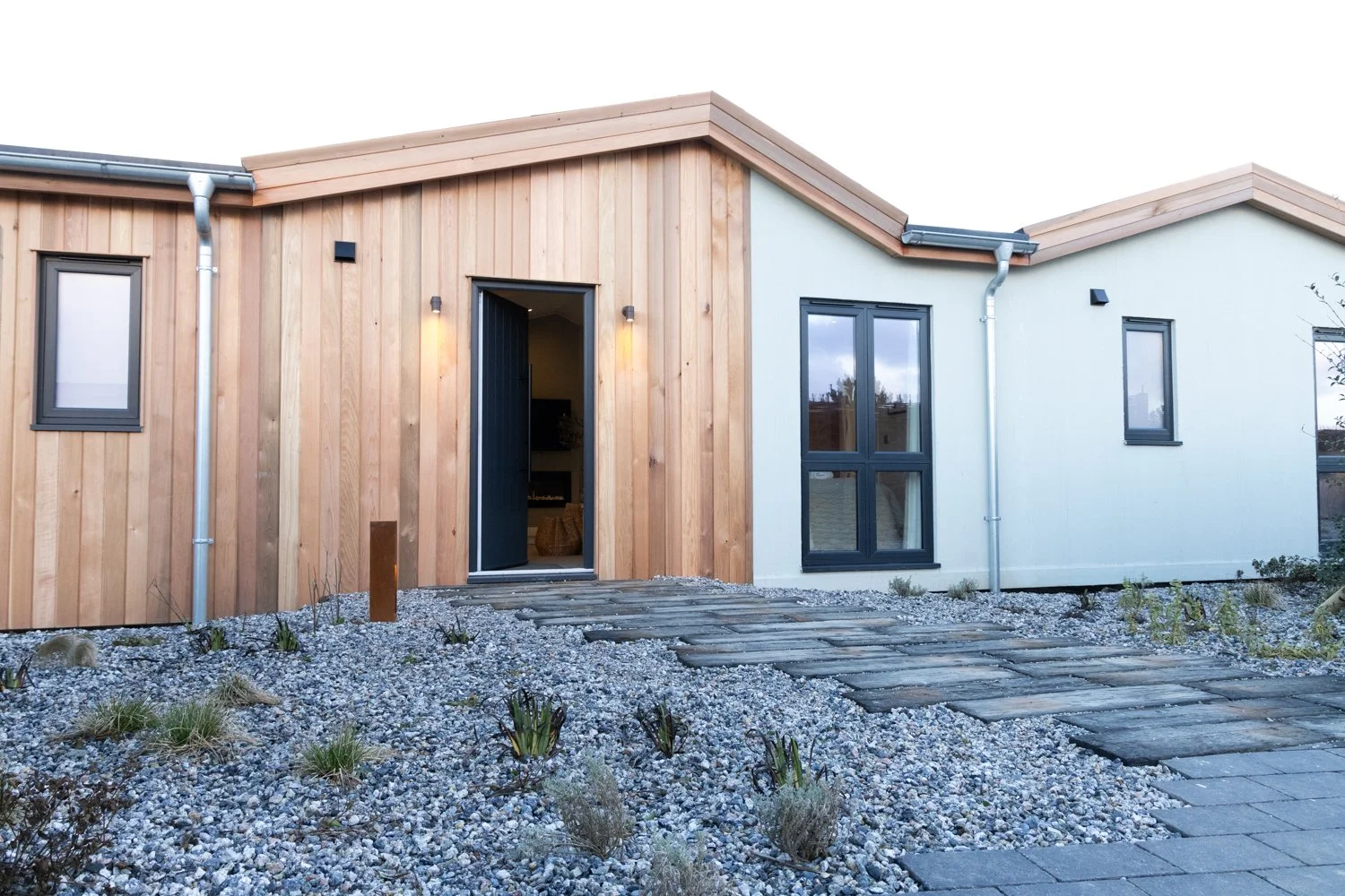 Modern house exterior with wood and light-colored siding, multiple windows, pathway with stone slabs, and gravel landscaping with small plants.