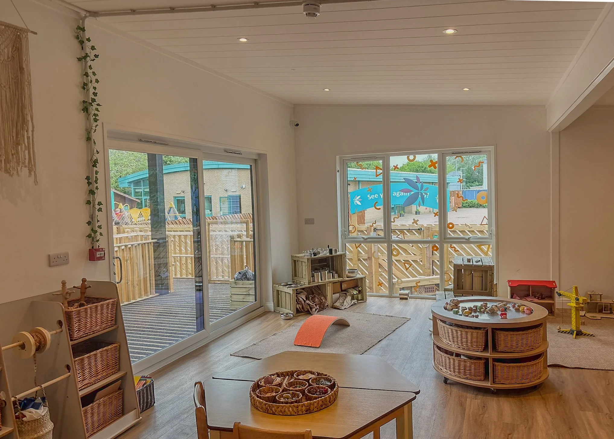 A bright children's playroom with large glass sliding doors leading to an outdoor area with wooden fencing and colorful structures. Inside, there are wicker baskets, small tables, chairs, and various toys, with a white ceiling and wooden flooring.
