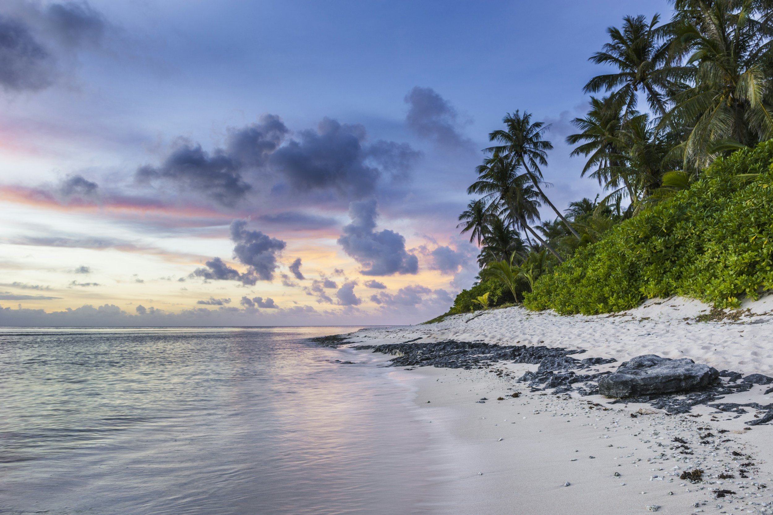 A tropical beach at sunset with white sand, black rocks, lush green palm trees, and a colorful sky with clouds.