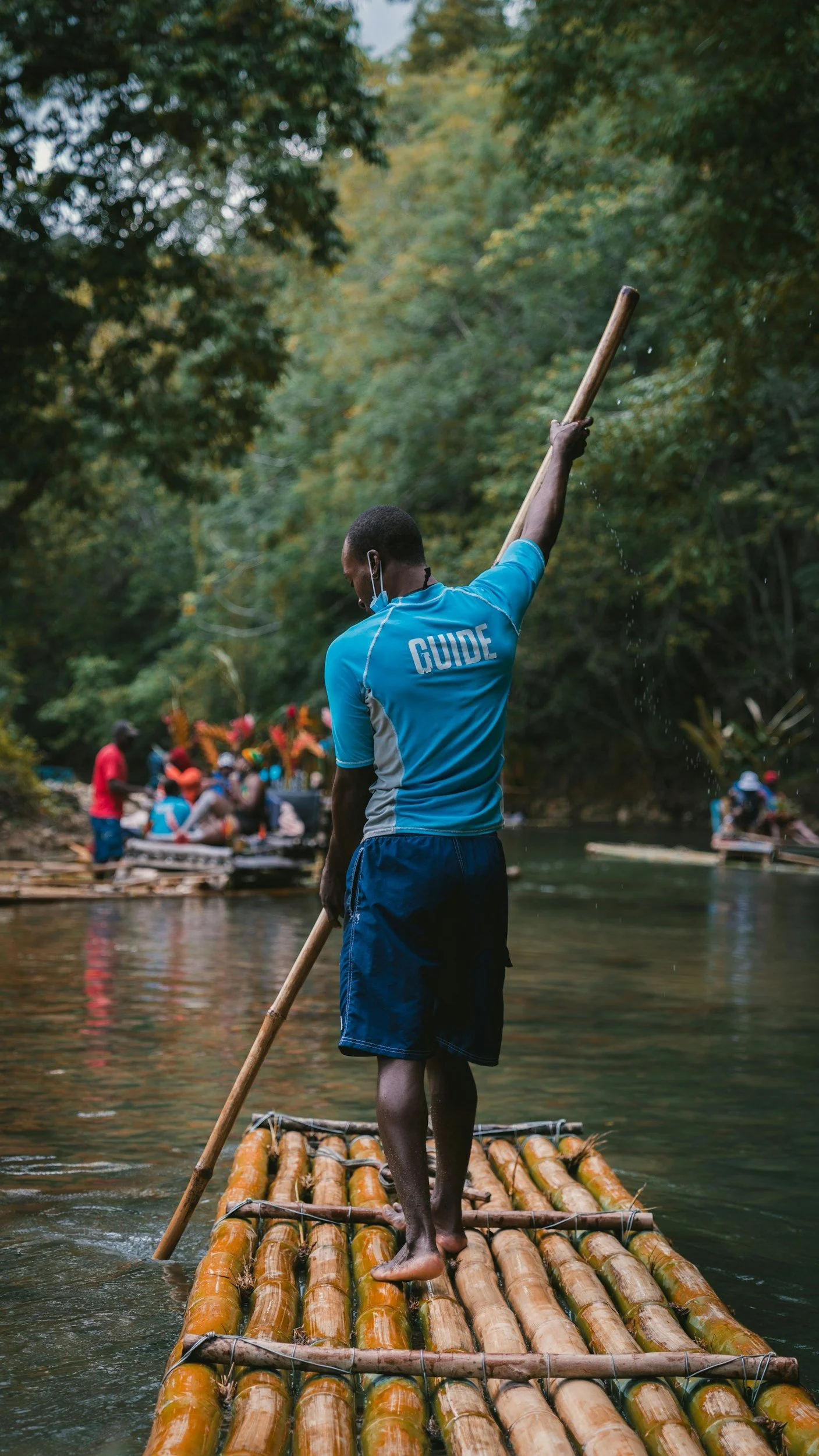 A guide standing on a bamboo raft on a river, holding a paddle, with a group of people on another raft in the background surrounded by lush green trees.