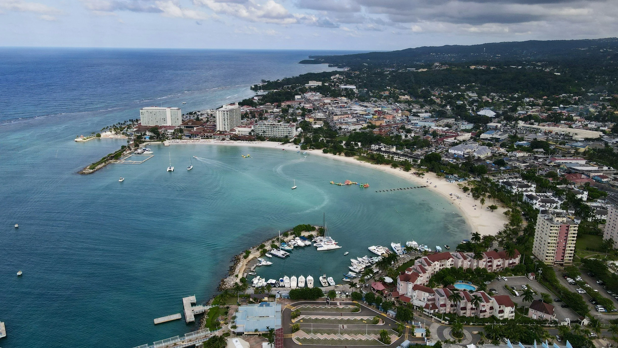 Aerial view of a crescent-shaped beach with white sand, turquoise water, boats, and yachts docked at a marina. Surrounding the beach are hotel buildings, residential apartments, and a cityscape with numerous small buildings. The ocean extends into the horizon with a cloudy sky overhead.