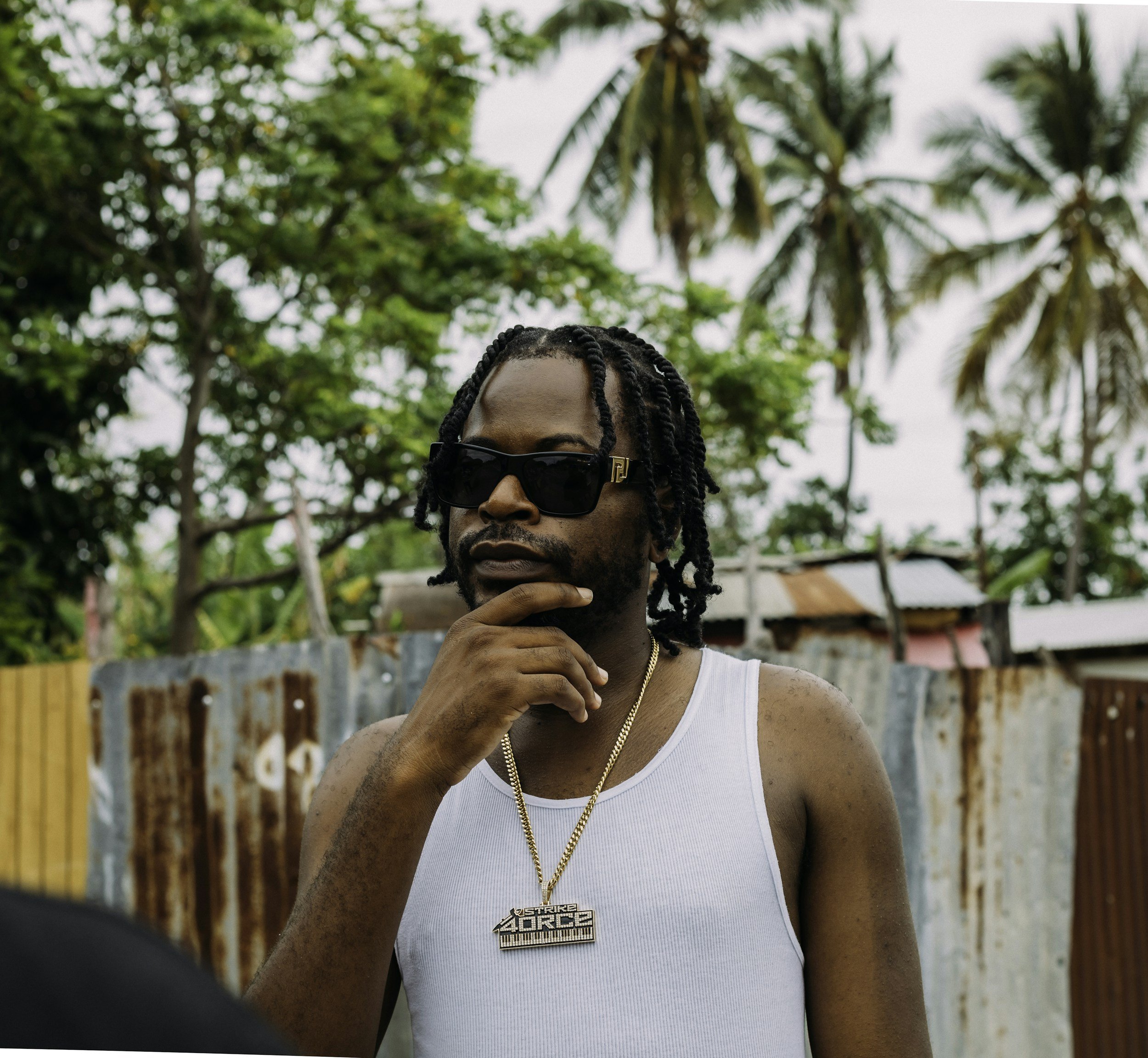 A man with dreadlocks, wearing sunglasses, a white tank top, and a gold chain necklace with a pendant that reads 'Strike Force,' posing outdoors with a thoughtful expression.