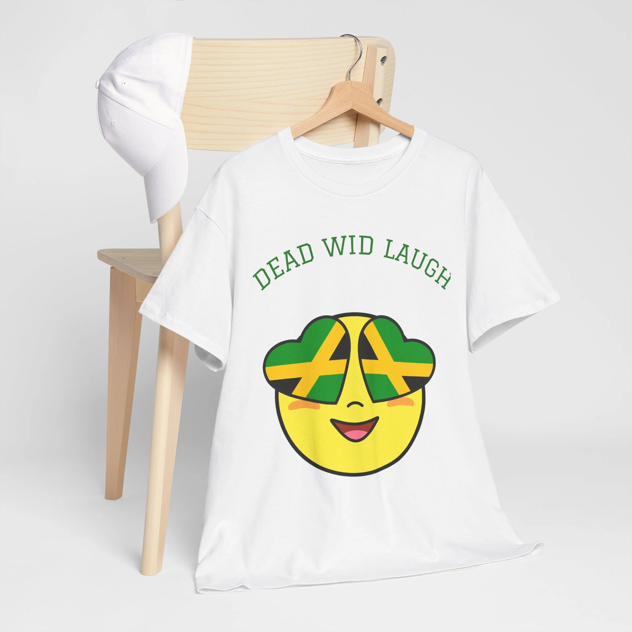 White T-shirt with a cartoon smiley face wearing sunglasses that have the Jamaican flag pattern, and the words 'DEAD WID LAUGH' above the face. A white cap hangs on a wooden coat rack in the background.