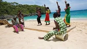 Group of people dancing and performing on a sandy beach with the ocean in the background.