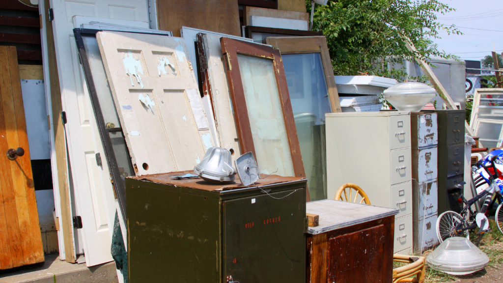 Outdoor scene with various old furniture and appliances, including filing cabinets, doors, a wooden chair, a bicycle, and lamps, stacked or standing among greenery.