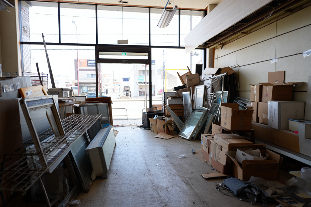 Interior of a store or storage room with disorganized boxes, frames, glass panels, and miscellaneous items scattered around, with large front windows allowing natural light in.